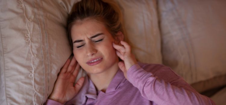 Woman lying in bed holding her jaw and ears, showing discomfort from nighttime teeth grinding