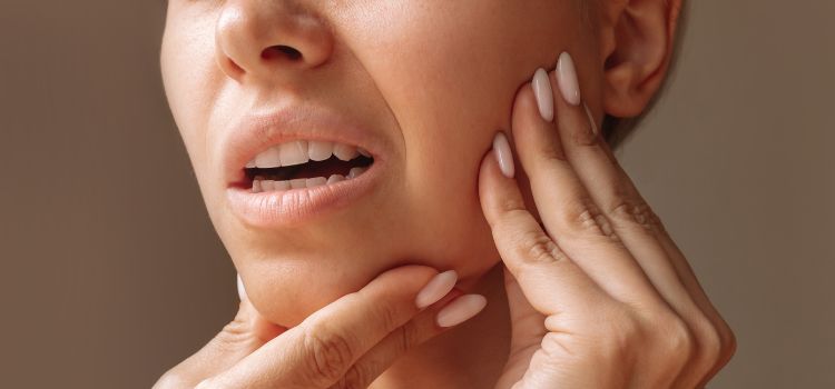 Close-up of a young woman touching her jaw with both hands, appearing to experience jaw pain or discomfort, possibly due to bruxism or TMJ issues.