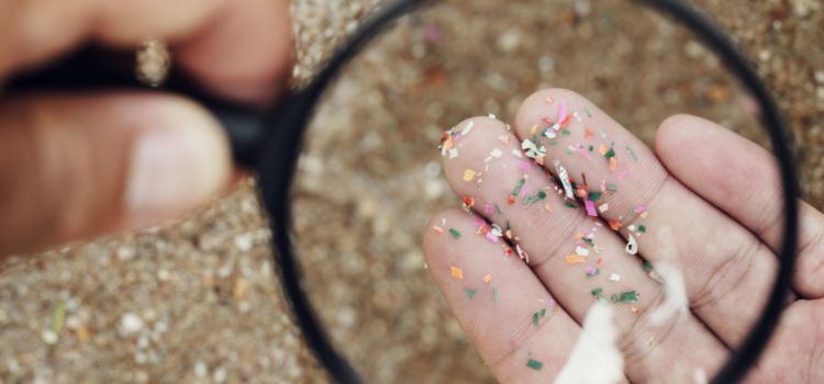 A hand holding microplastic particles under a magnifying glass on a sandy surface.