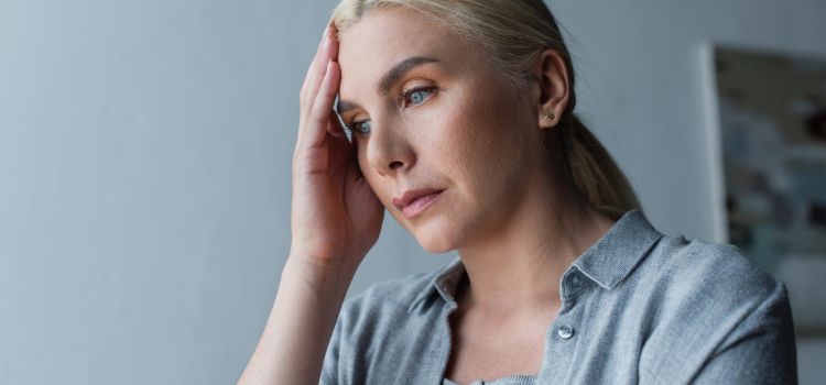 Blonde woman touching her forehead, appearing stressed and tired in a gray shirt